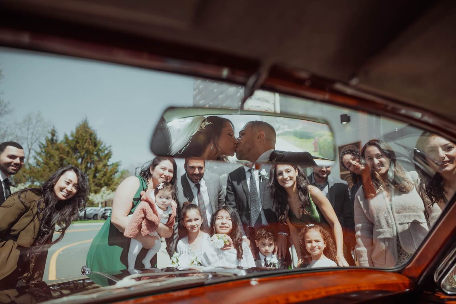 Christina and Mike kiss reflected in the vintage car window as family gathers around, Holmdel NJ