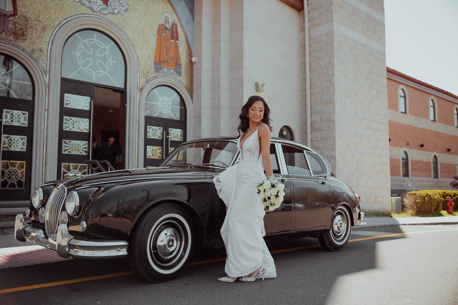 Bride in her gown leaning on a vintage car at the doors of St. Mina Coptic Orthodox Church