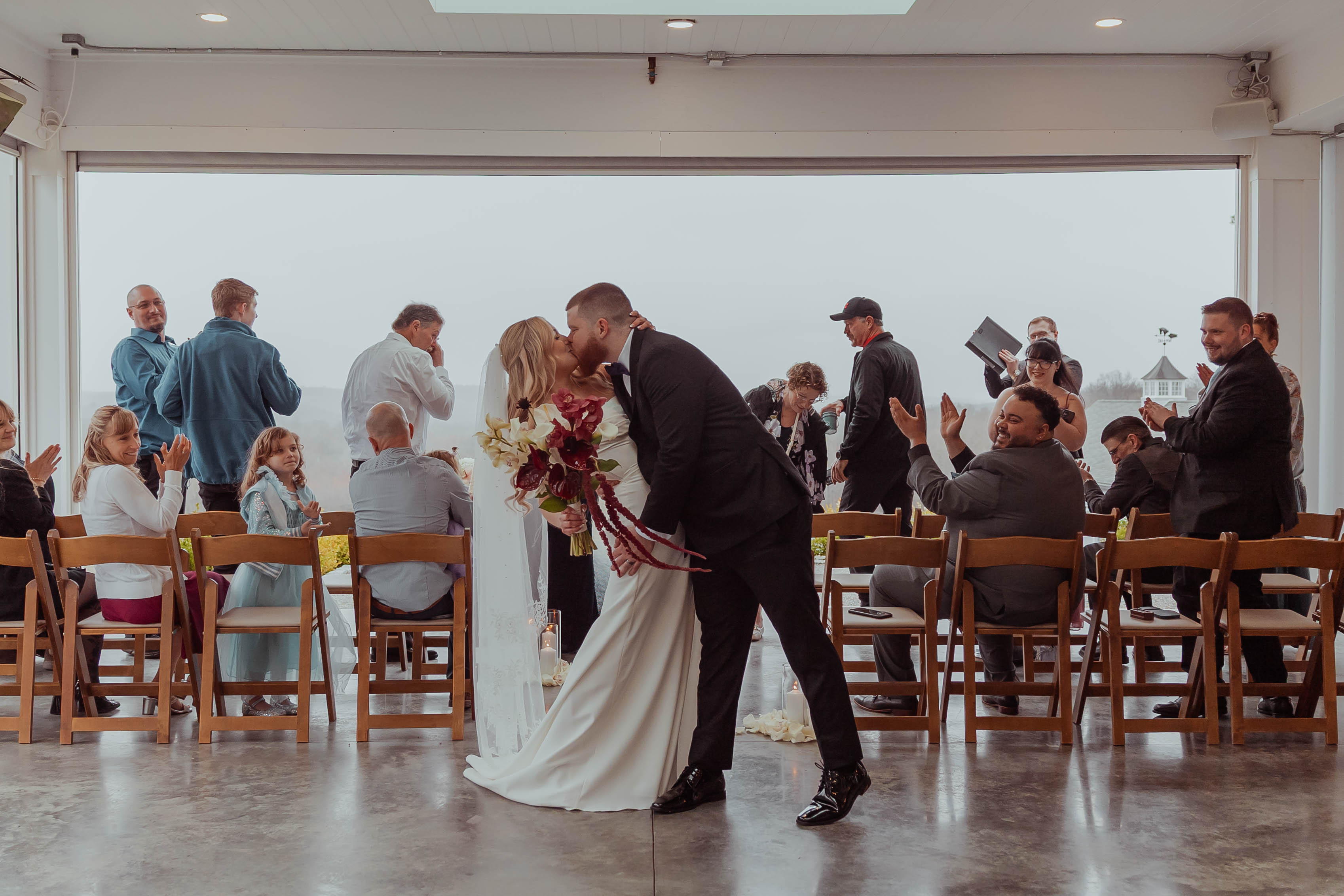 Rebecca and David kiss after their ceremony in the glass room at Milea Estate Vineyard, Staatsburg NY, with a foggy vineyard behind them
