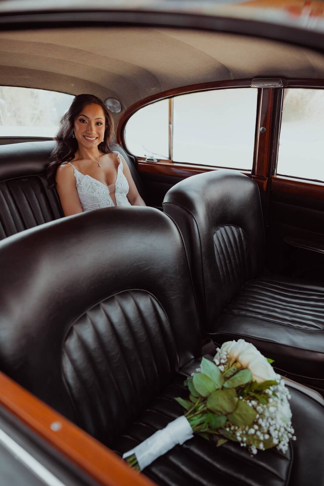 Bride seated in a vintage car with her white rose bouquet resting on the leather seat