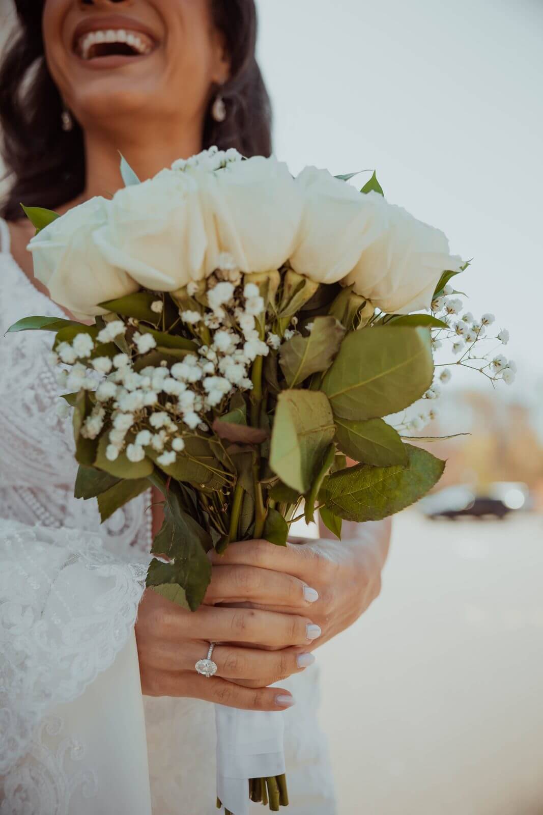 Bride laughing while holding a white rose and baby's-breath bouquet, engagement ring visible